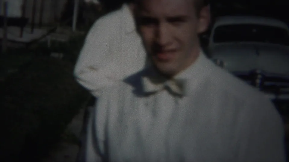 A man in a white shirt and bow tie stands close to the camera, with a mid-century car visible behind him in Westville.