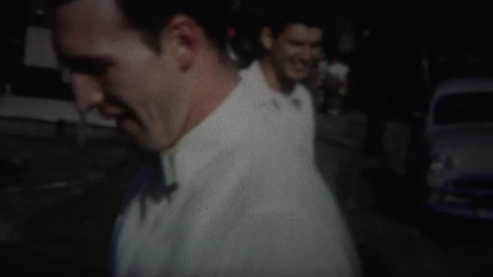 A smiling man passes near the camera while another attendee appears behind him in the dim 1951 reunion footage.