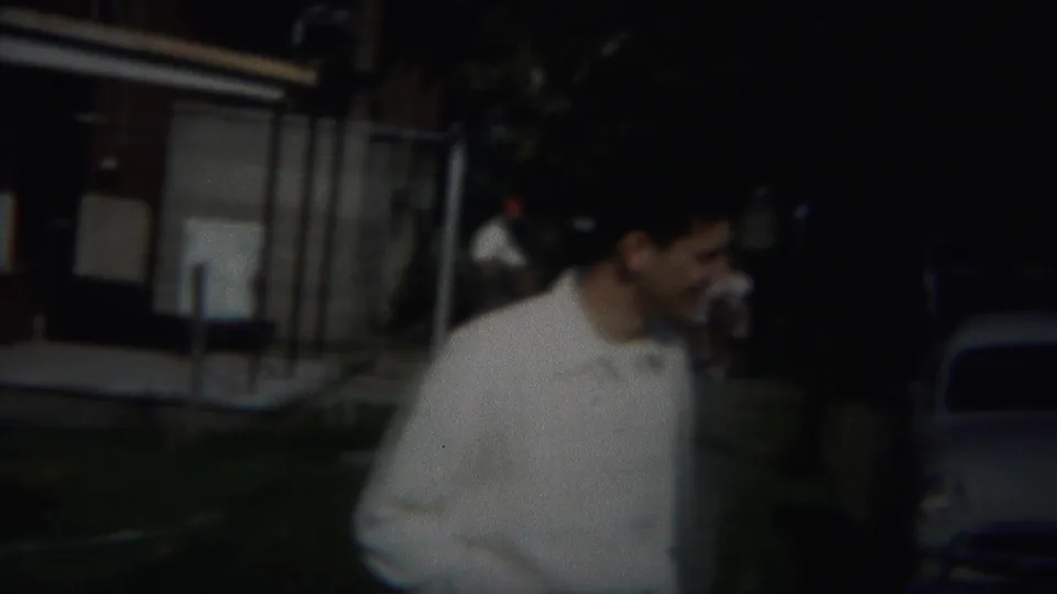 A bow-tied man walks through a shadowed outdoor residential area, with a parked 1950s-style car behind him.