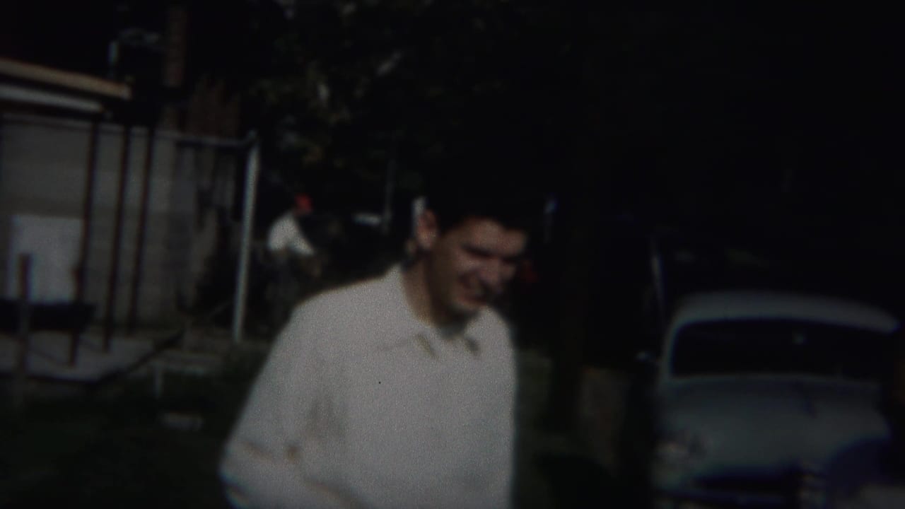 Man in a white shirt stands near a shaded street with parked cars behind him.
