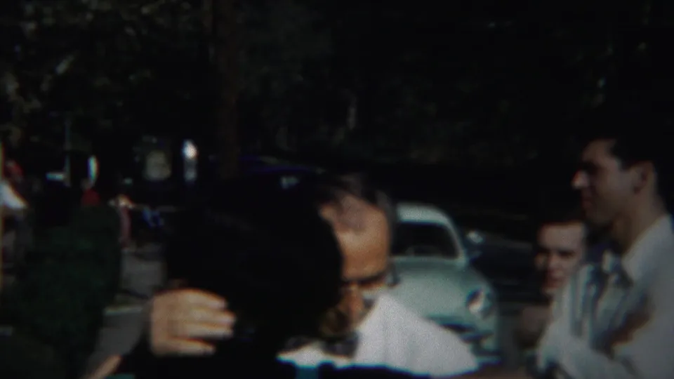 A dark-haired young woman smiles close to the camera while adults in white shirts stand nearby in the Westville home-movie scene.
