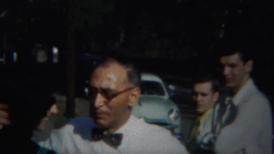 An older man wearing glasses and a bow tie moves past the camera, with men in white shirts and light-colored cars behind him.