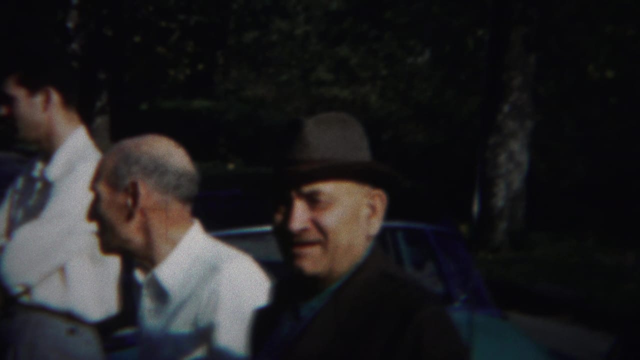 Older man in a fedora looks toward the camera while two men stand nearby.