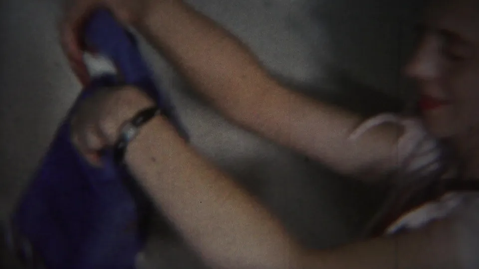 In supplied 1951 Westville home-movie footage, a woman in a dark dress lifts a blue garment in a softly lit interior.