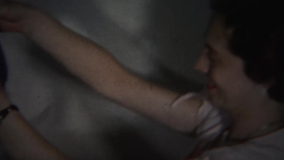 A smiling woman, wearing earrings and a dark dress, holds up a blue garment in the 1951 Westville home-movie scene.