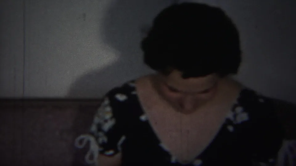 The woman in a black floral dress looks down in a dim private interior, with upholstered seating and a plain wall behind her.