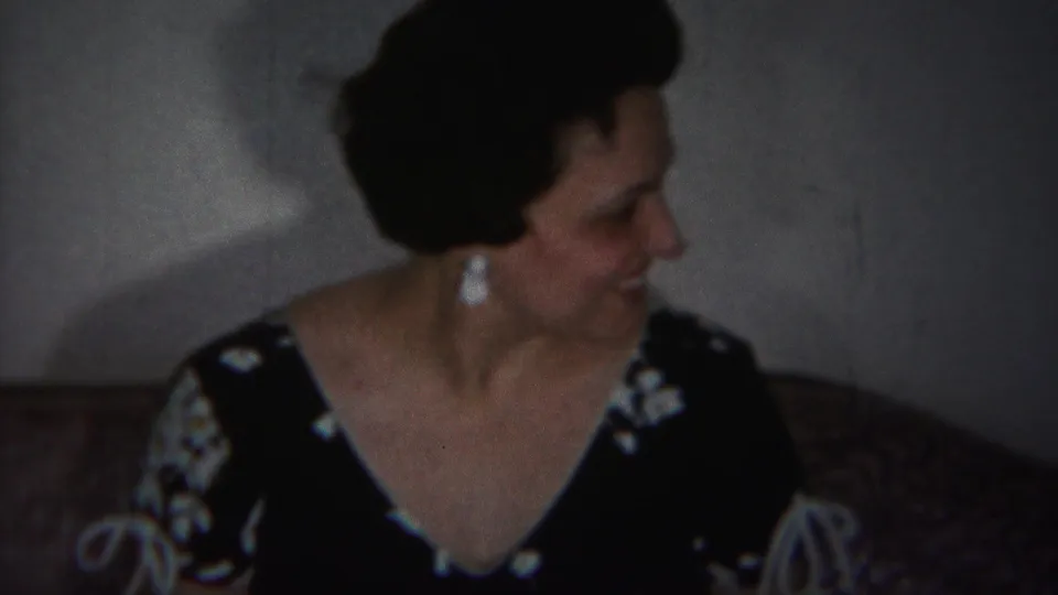 In 1951 Westville, New Jersey home-movie footage, a woman in a dark floral dress and pearl earrings smiles on a couch.