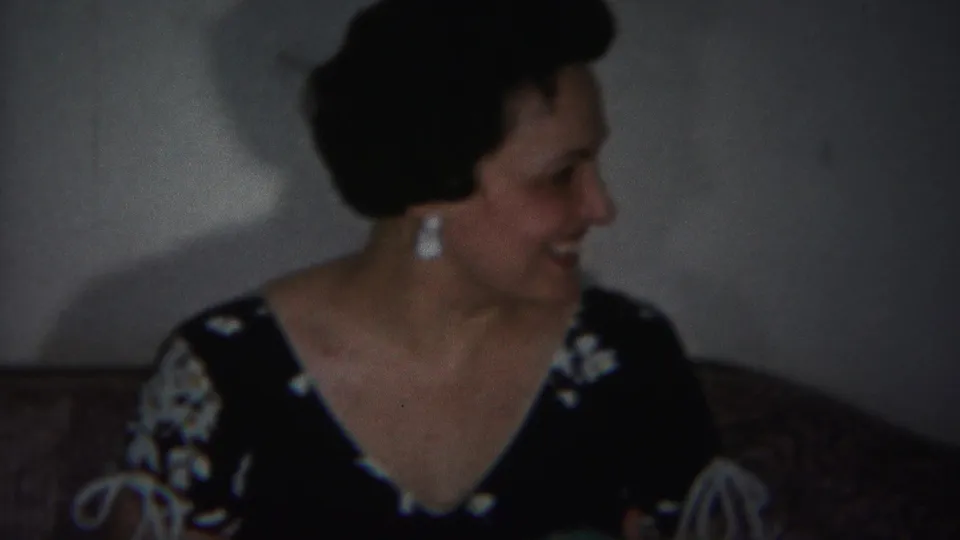 The woman remains seated on the patterned couch, turned right as if listening or responding during a 1951 home-movie moment.