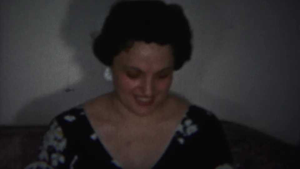 In the dim interior, the woman looks down with a smile, her dark updo, earrings, and floral dress visible.