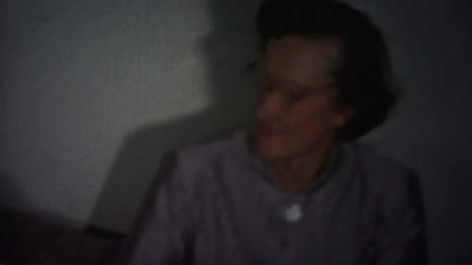 In supported 1951 Westville, New Jersey home-movie footage, an older woman with glasses sits indoors before a plain wall in dim light.