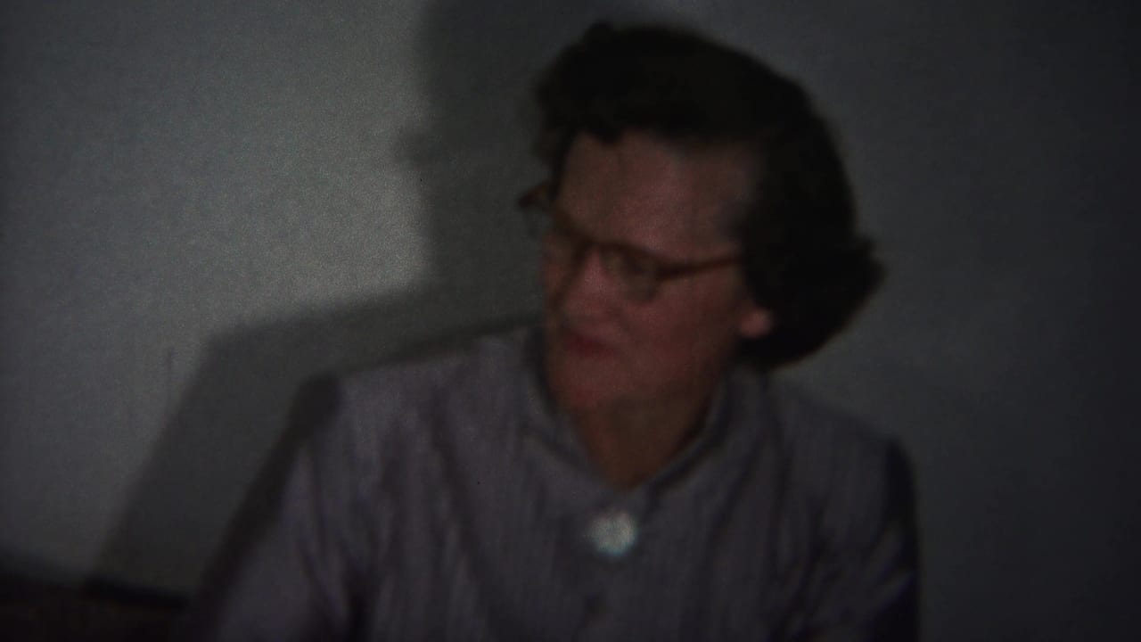 Older woman with glasses facing left in a dim room with a strong wall shadow behind her.