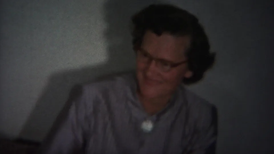 In the 1951 home-movie frame, the older woman smiles while a second person is partly visible nearby in the dim private room.