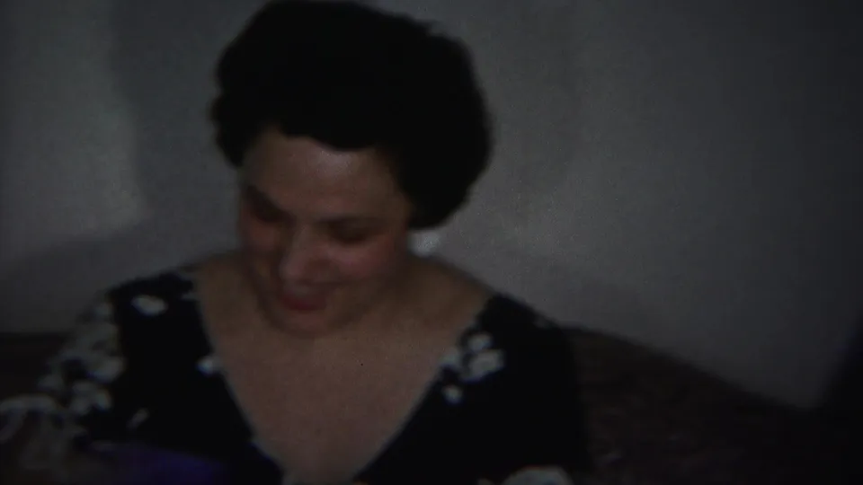In a 1951 Westville home-movie frame, two women sit indoors on upholstered seating, with one woman looking down in a dark dress.