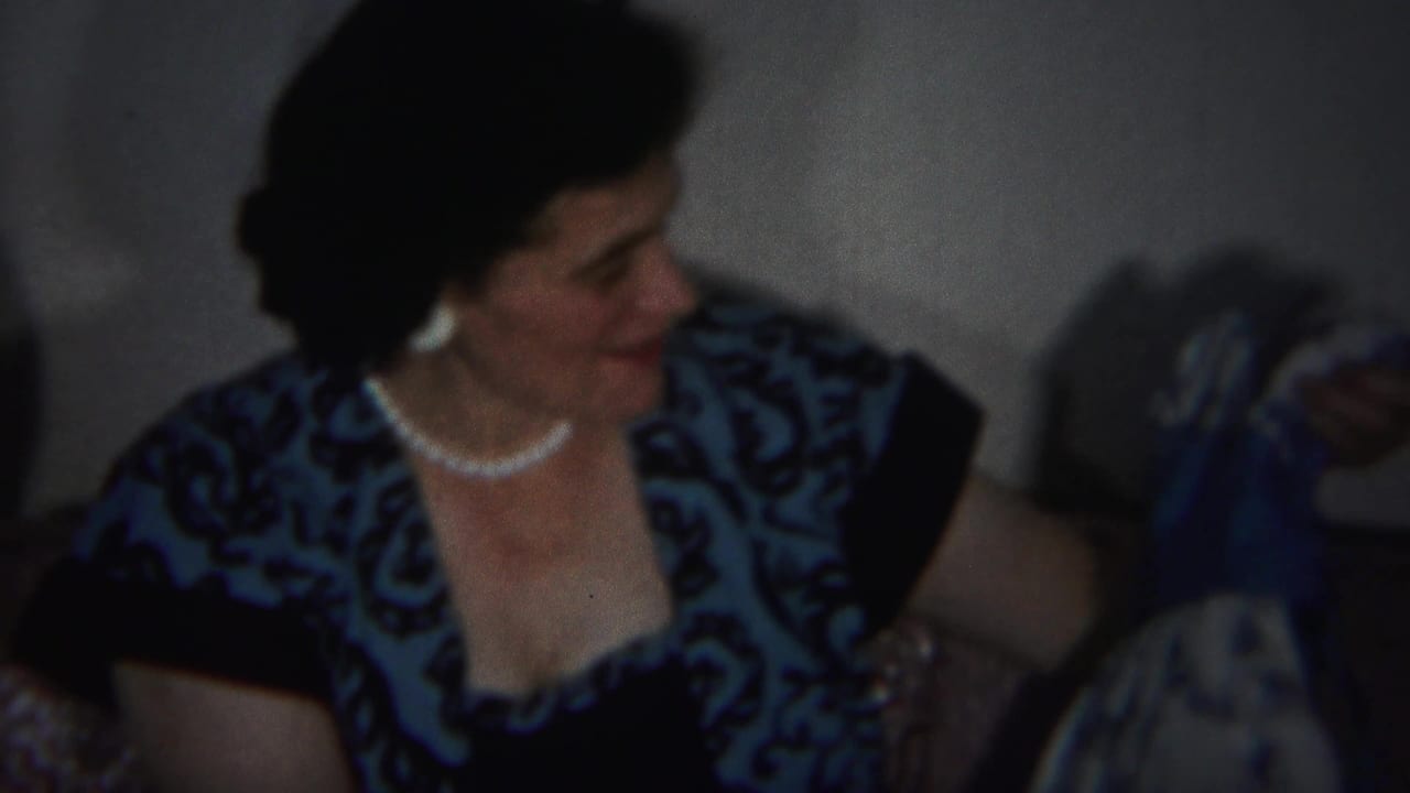 Woman in a blue patterned dress and pearl necklace turns while seated indoors.