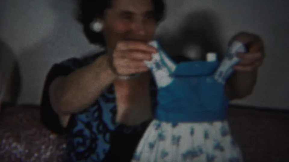 The woman smiles as she lifts a small blue-and-white baby dress for the camera in the 1951 Westville home-movie scene.