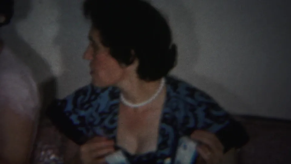 A woman in a pearl necklace sits indoors beside another person, holding part of the baby dress in 1951 Westville footage.