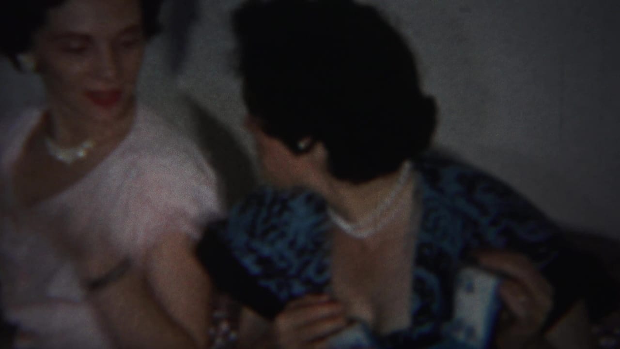 Two women seated indoors, one in a pale pink dress and pearls and one in a dark patterned dress.