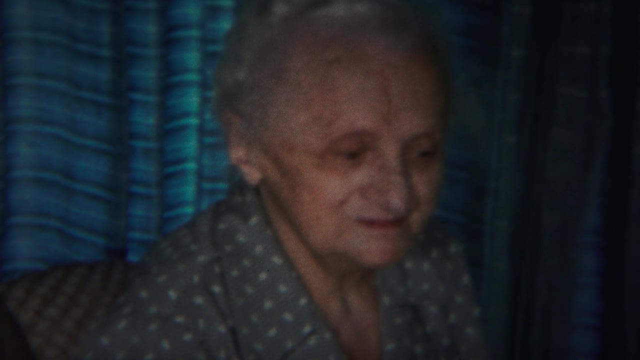 Elderly woman in a gray polka-dot blouse seated near blue striped drapes.