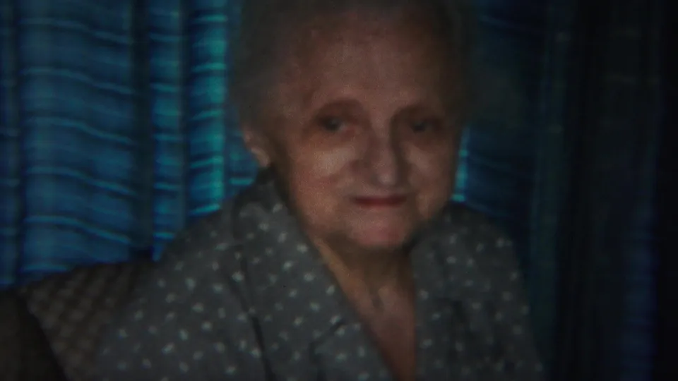 The elderly woman faces the camera more directly, wearing a gray polka-dot blouse against blue striped drapes in the indoor 1951 footage.