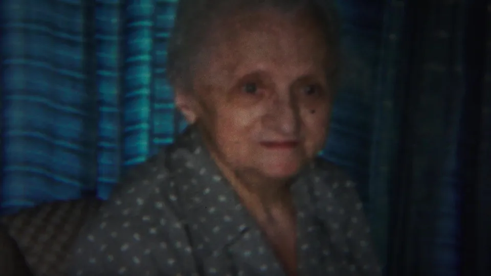 The elderly woman remains seated indoors, framed by blue striped drapes and nearby patterned upholstery in the Westville home-movie clip.