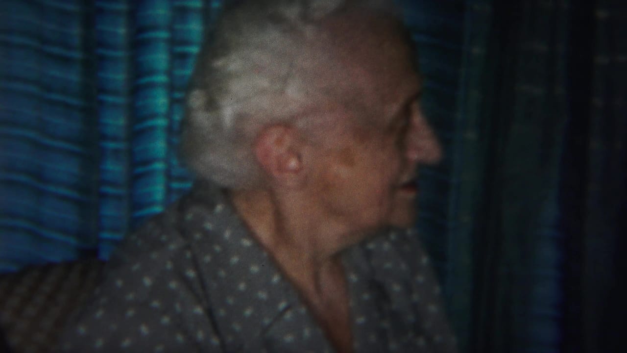 Elderly woman in a polka-dot blouse seated beside blue curtains.