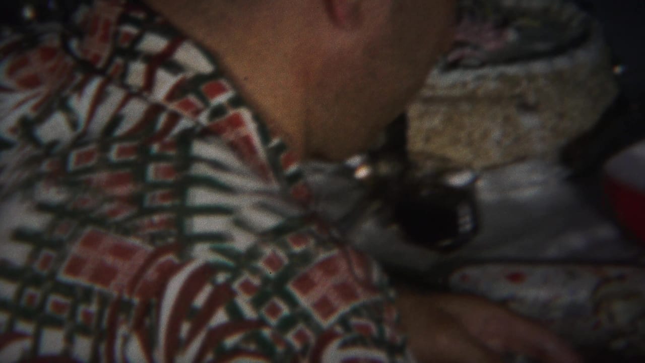 Adult man in a patterned shirt seated in a dim room.