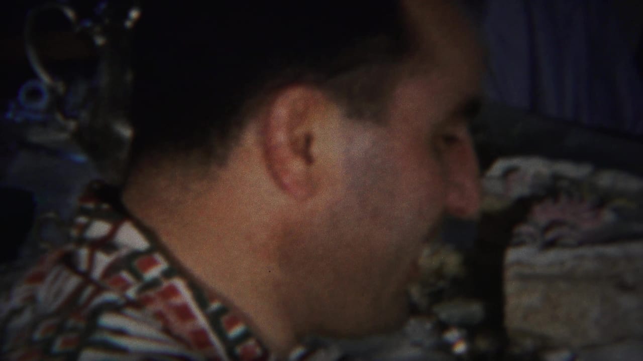 Man in a patterned shirt turns toward someone beside a table in a dim interior.