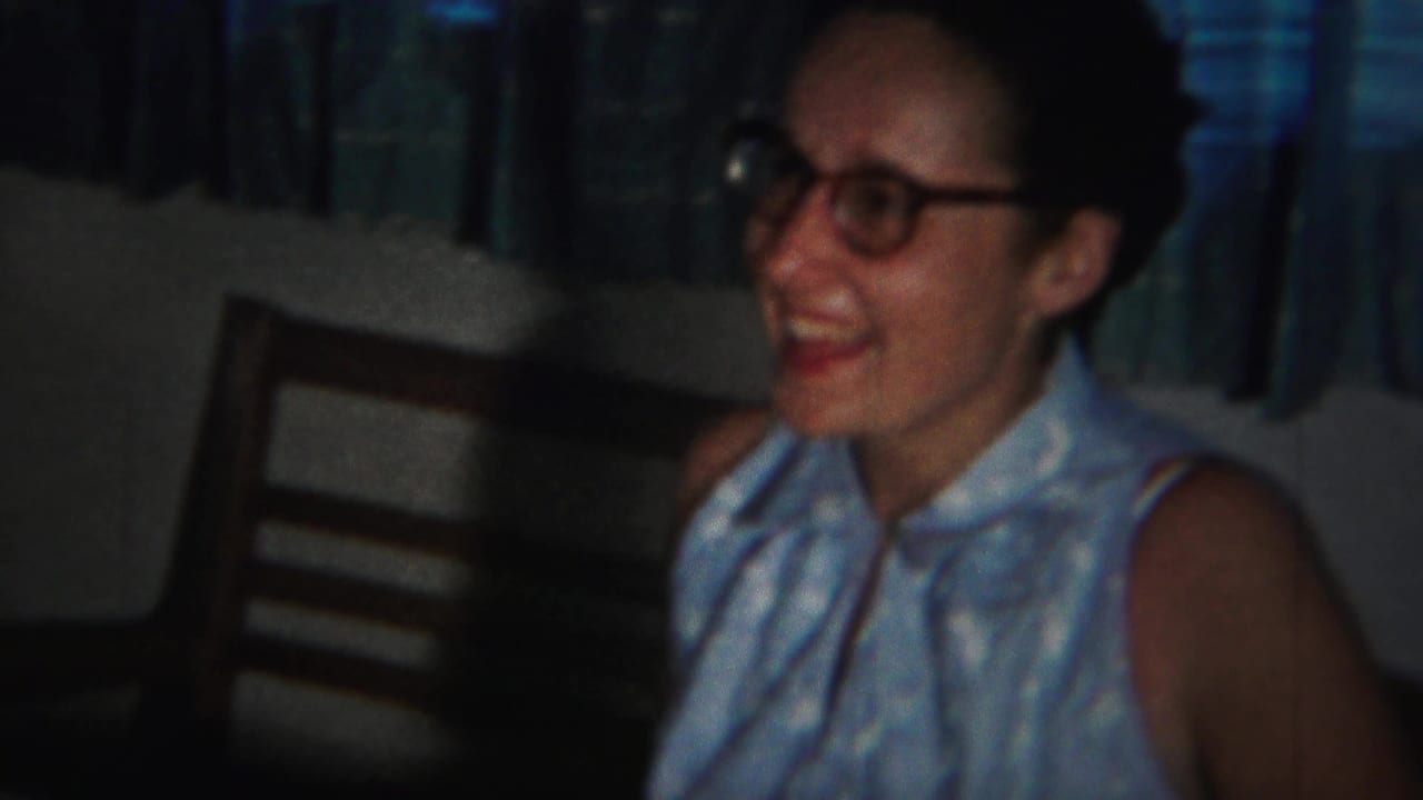 Smiling woman in glasses and a light sleeveless dress seated indoors near curtains.