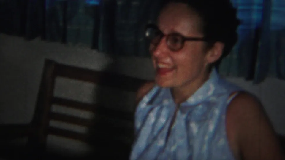 In a 1951 Westville, New Jersey home-movie frame, a woman in glasses smiles beside patterned curtains in a dim interior.