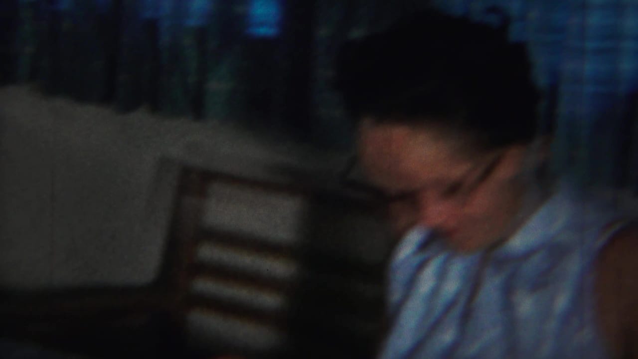 Woman in glasses looks downward beside a wooden chair and blue curtains.