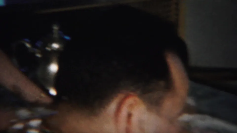 A blurred close indoor frame shows part of an adult at a table with a reflective object in a private home setting.