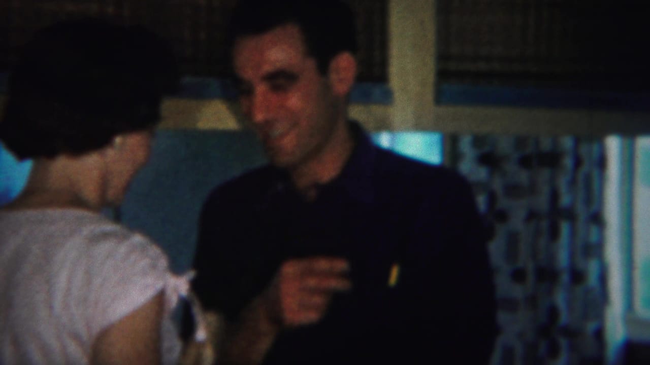 Man in a dark shirt talks with a woman seen from behind in a dim home interior.