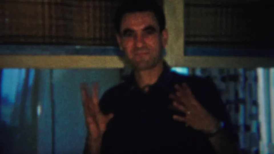 The man faces forward with both hands lifted mid-gesture, framed by curtains and a woven shade in the 1951 New Jersey home movie.