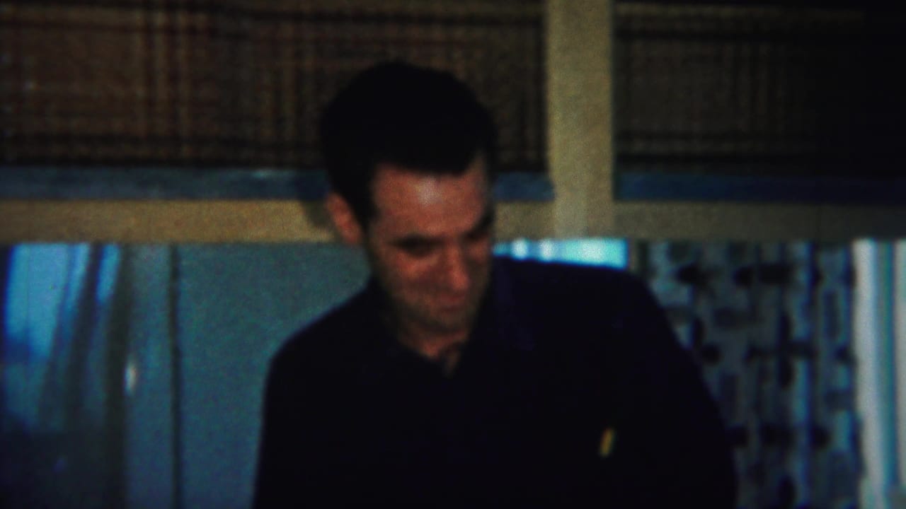 Man in a black shirt sits at a dim kitchen table with another adult partly visible nearby.