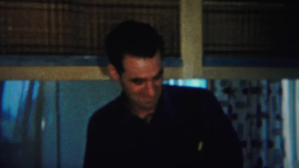 In 1951 Westville home-movie footage, a man in a dark shirt sits in a dim kitchen near shelves and patterned curtains.