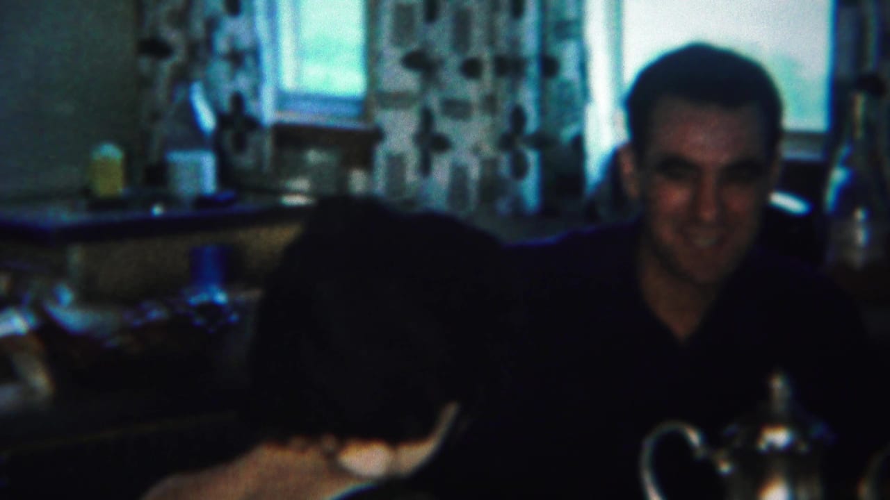 Man in a dark shirt sits beside a woman at a kitchen table with a teapot in front of them.