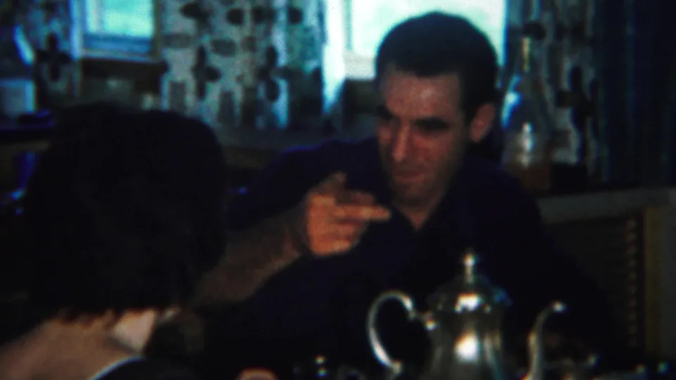 The man gestures while speaking across a kitchen table, with the woman partly turned away and a silver teapot visible below.
