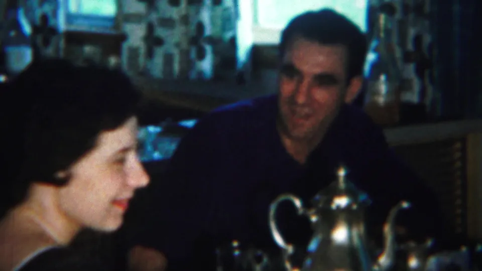 A woman smiles in profile beside a silver teapot while the man across the table smiles back in the 1951 home interior footage.