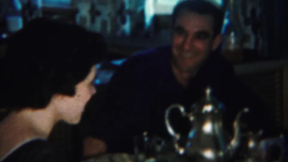 The woman turns toward the seated man at the dinner table, with a silver teapot and glassware in the foreground.