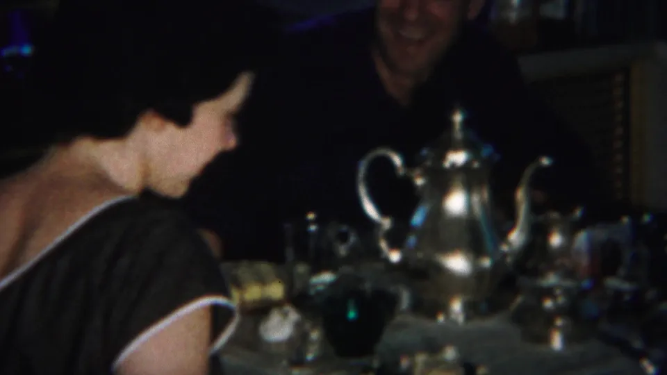 A side view shows the woman facing the man across a dimly lit table, with the silver teapot prominent in front.