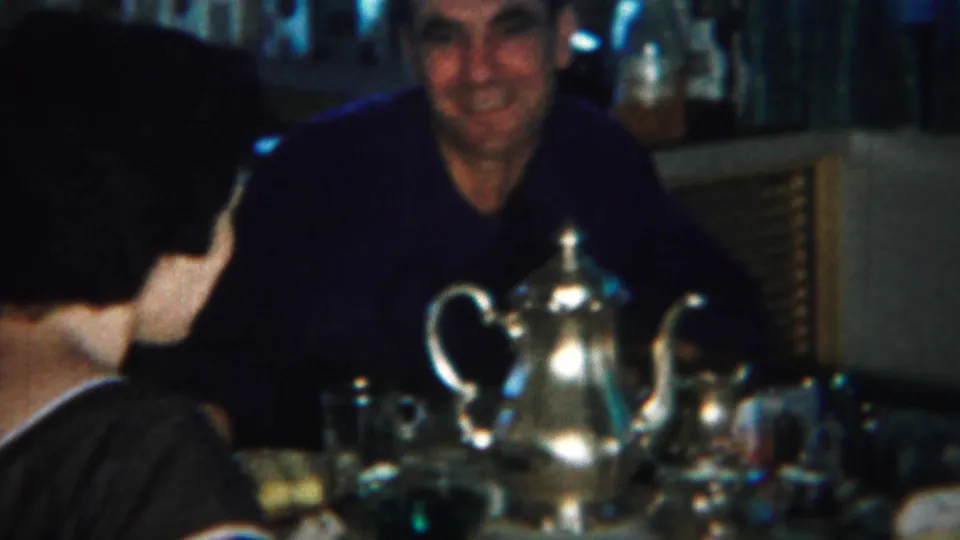 The man smiles across the table as the woman is seen from behind, with the silver teapot and glassware between them.