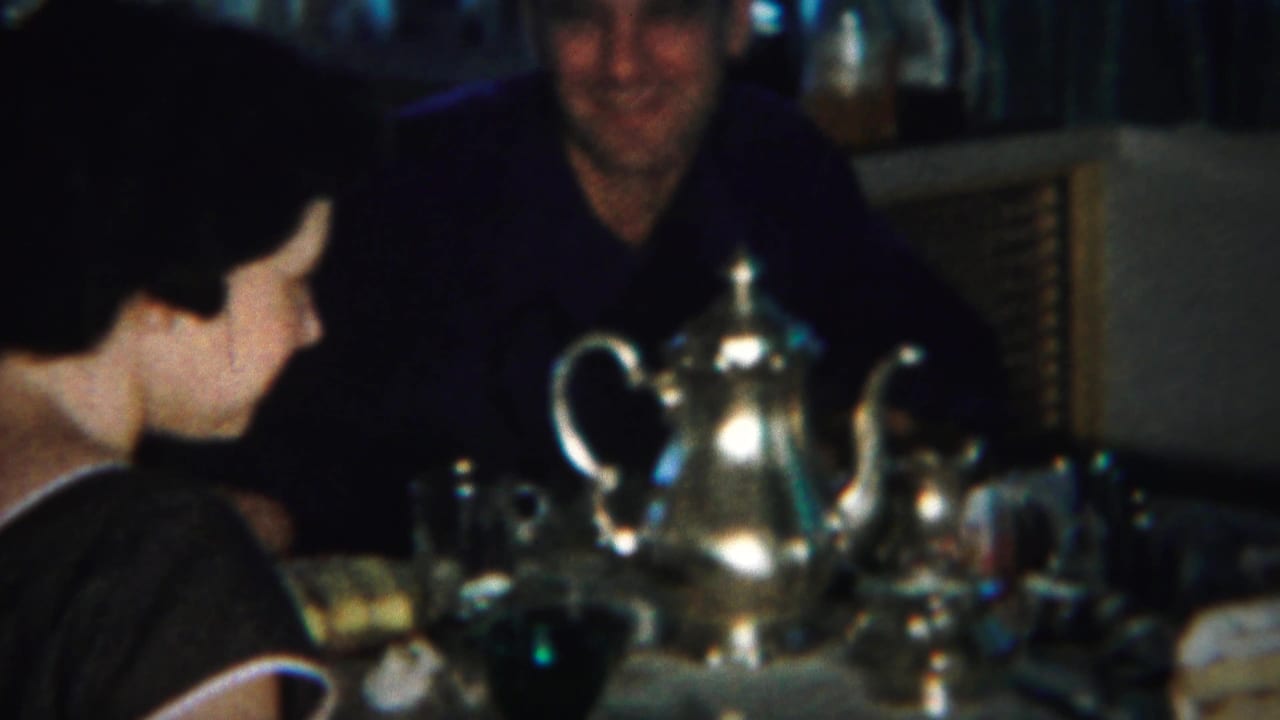 Three adults seated around a dim dining table with cake, cups, and a silver teapot.