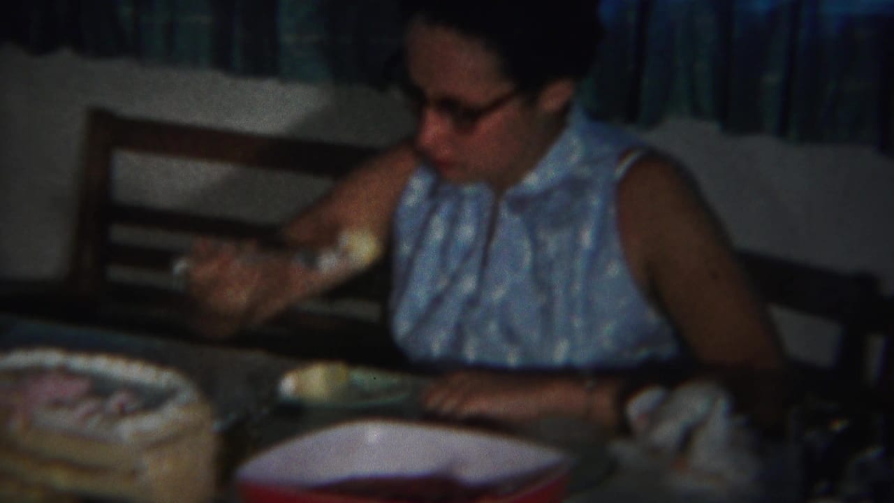 Woman in glasses and a blue sleeveless dress sits at a table with cake and dishes.
