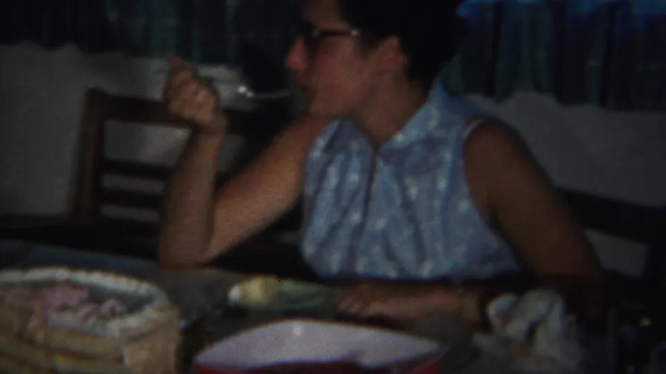 In the 1951 Westville home movie, the woman lifts a fork near her mouth while seated at a cake-covered dining table.