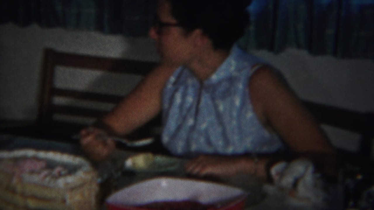 Adult woman in glasses sits at a table beside a decorated cake and dishes.