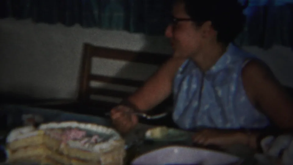 An adult woman in glasses and a blue sleeveless dress sits beside a decorated cake in the 1951 home-movie scene.