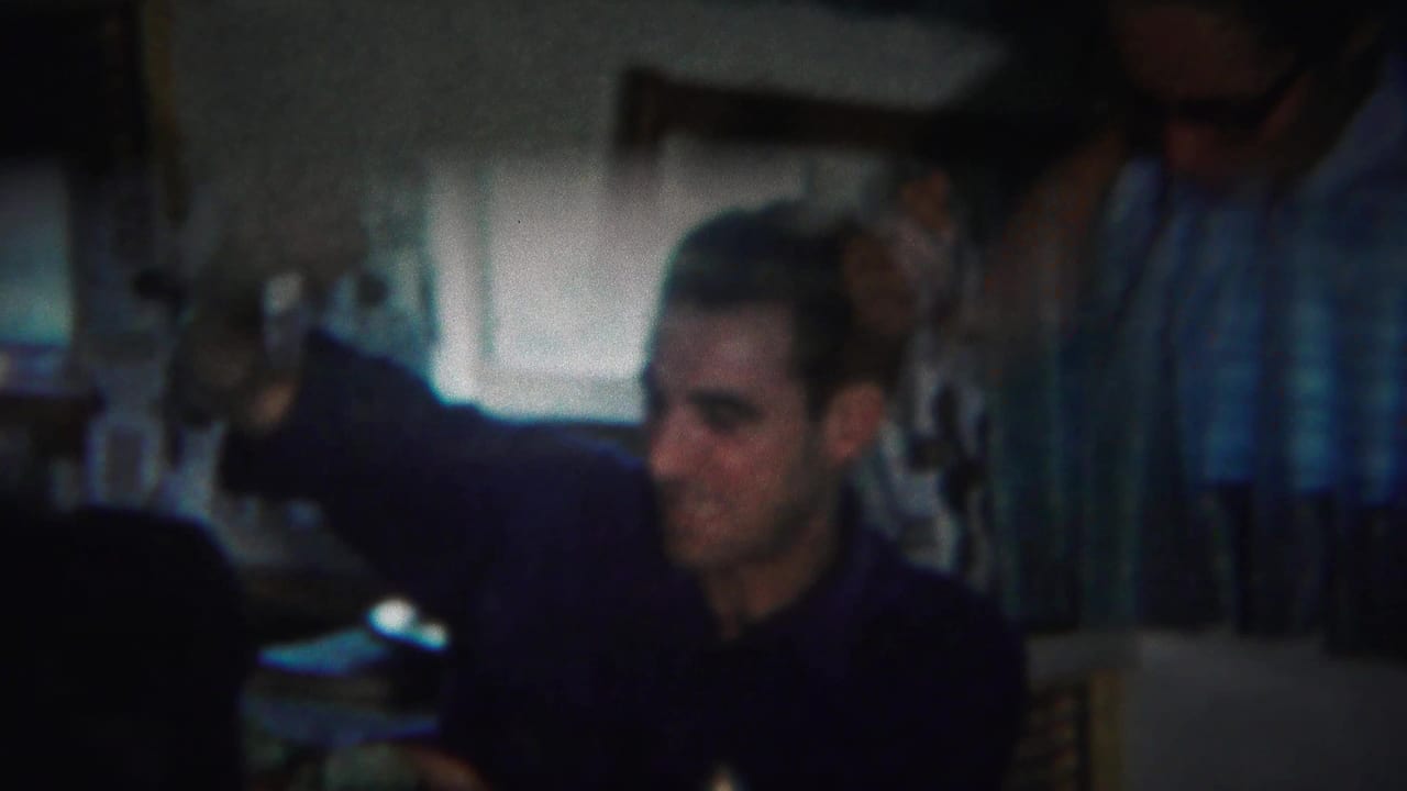 Two adult men indoors near a table, with one man partly visible behind another.