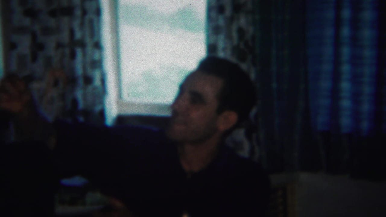 Adult man in a dark shirt raises one hand inside a room with curtains and a window.