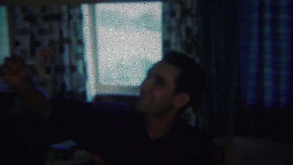 1951 home-movie frame of a man gesturing upward in a private Westville interior with curtains and window light behind him.