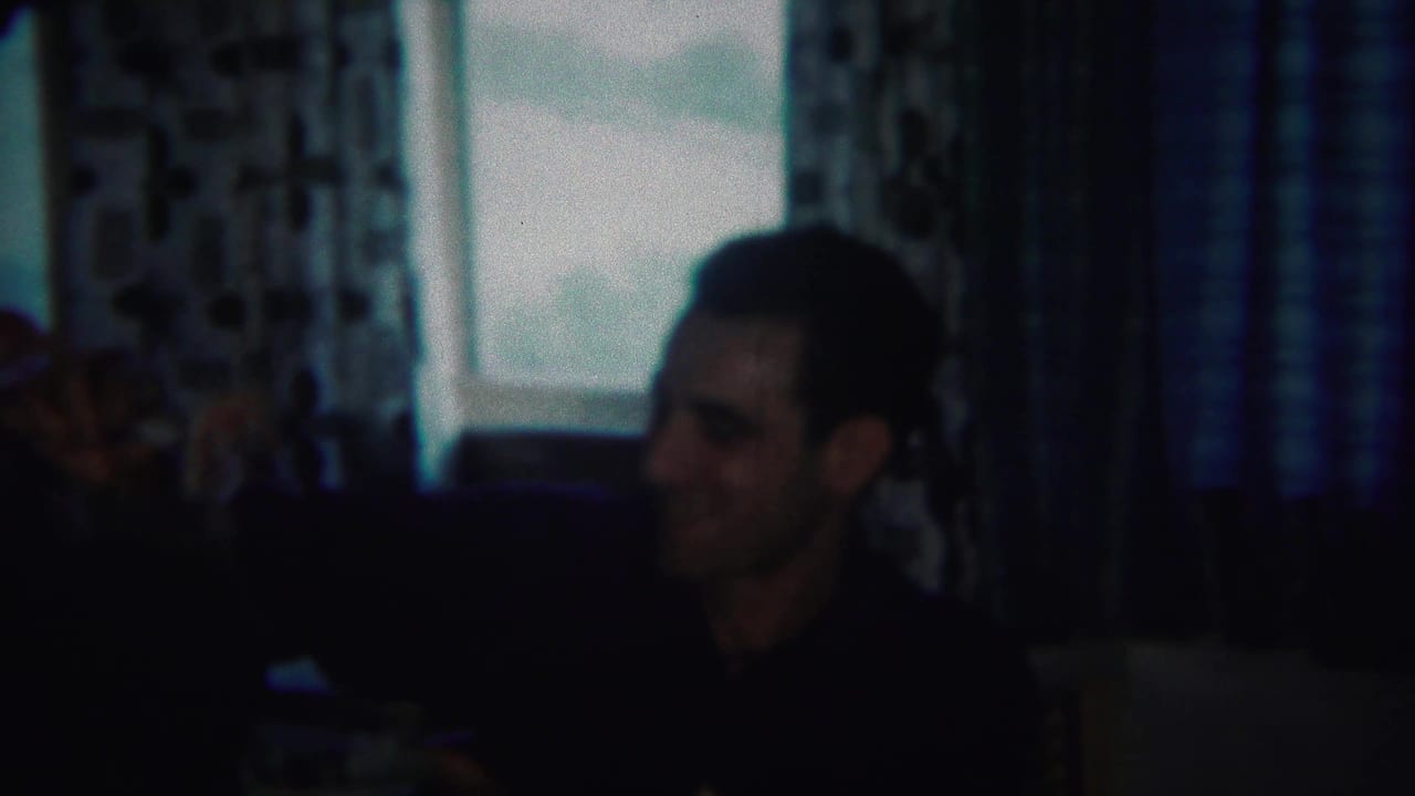Man seated in a dim room with patterned curtains and bright windows behind him.
